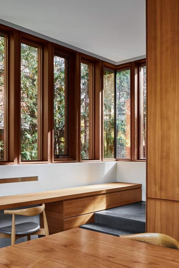 A warm, wood-lined interior corner with a dining table in the foreground, a built-in timber bench beneath a continuous bank of tall windows, and dark stepped flooring leading toward the window seat. The extensive glazing overlooks leafy trees, while white walls and ceiling provide contrast to the rich timber joinery.