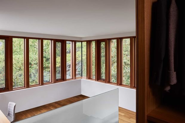 A bright, modern interior corner with extensive floor-to-ceiling timber-framed glazing wrapping around two walls, overlooking dense greenery. White walls and a white balustrade or partition contrast with warm timber flooring and dark built-in timber joinery at the right. A small portion of a white chair is visible in the foreground.