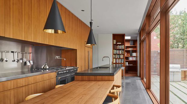 A contemporary galley-style kitchen with extensive warm timber cabinetry, stainless-steel splashback and appliances, dark stone benchtops, a central island, and a long timber dining/work table. Black pendant lights hang from a white ceiling. Full-height timber-framed glazing lines one side, opening views toward a paved courtyard, while built-in timber bookshelves occupy the rear wall.