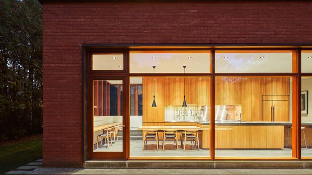 Exterior view of a contemporary brick building with a warmly illuminated kitchen and dining area visible through expansive timber-framed glazing. The interior features vertical timber wall panelling, a long kitchen island with seating, pendant lights, and a dining table. Dark red brick cladding forms the solid facade around the glazed opening, with mature landscaping visible at the side.
