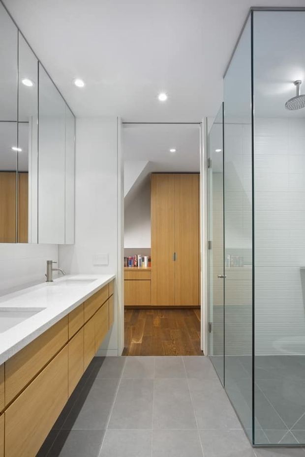 Bright contemporary bathroom viewed toward an adjoining dressing or wardrobe area. The room features a long white vanity with light timber drawers, mirrored wall-fronted storage, large grey floor tiles, a frameless glass shower enclosure with a round overhead shower, and white tiled shower walls. A timber wardrobe and dark timber flooring are visible through the open doorway.