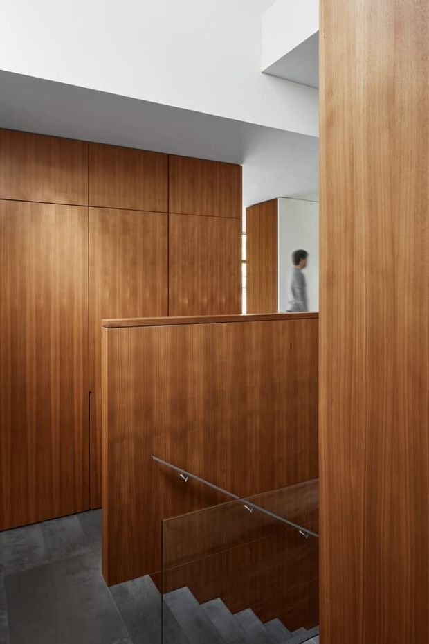 Interior view of a contemporary stairway with a descending flight of grey-finished steps, clear glass balustrade and slim metal handrail. The stair is enclosed by extensive warm timber wall panelling and built-in joinery, with a large mirror opening reflecting the space and a person. White angular ceiling planes create a crisp architectural contrast.