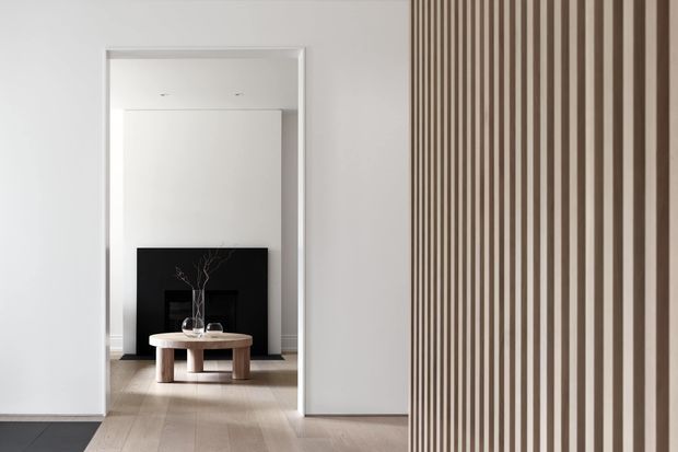 Minimalist interior view through a wide opening into a refined living room. Pale timber flooring, white walls and a large black fireplace feature create a strong monochrome composition, with a round timber coffee table and sculptural branch arrangement in front. A full-height vertical timber slat screen dominates the right foreground, adding rhythm and subtle separation between spaces.