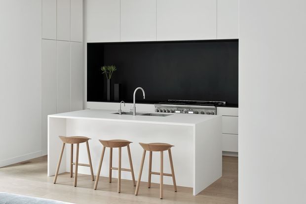 A bright, minimalist contemporary kitchen dominated by a large white island with an integrated sink and tall curved faucet. Three light timber bar stools line the island, while white full-height cabinetry surrounds a wide matte-black splashback and stainless-steel cooktop. Pale timber flooring and a restrained monochrome palette create a clean, refined interior.