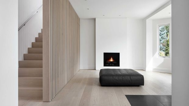 A bright minimalist living area with a pale timber staircase and matching vertical slatted screen, white walls, light timber flooring, a recessed fireplace, and a low black leather ottoman. A narrow window brings in greenery and daylight, while dark tile flooring appears at the right edge.