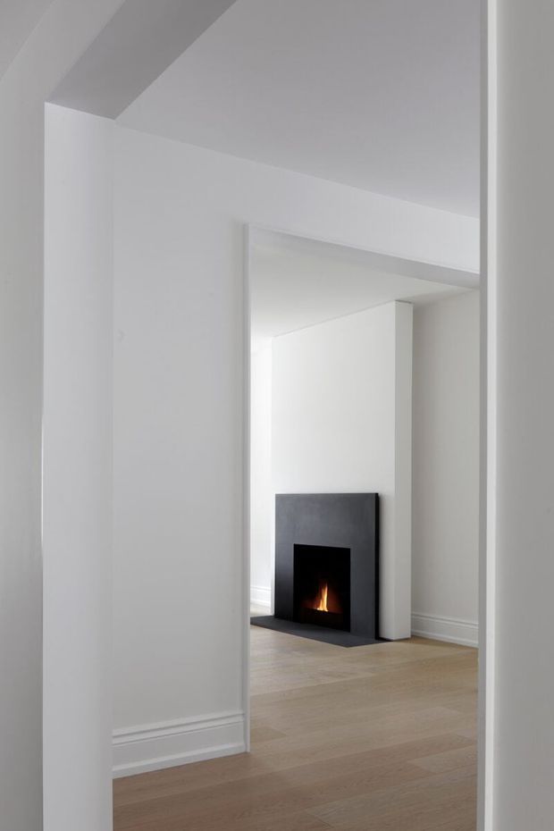 A minimalist white interior viewed through a doorway, showing an empty room with pale timber flooring, crisp white walls and trim, and a dark, simple fireplace with a small flame. The composition emphasizes clean architectural lines, recessed door openings, and restrained detailing.