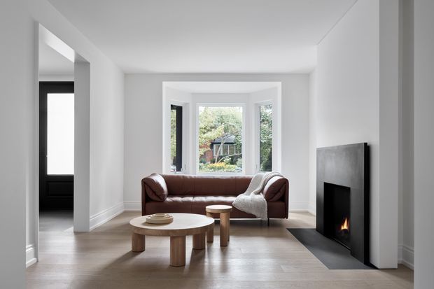 Bright, minimalist living room with white walls, pale timber flooring, a brown leather sofa, round timber coffee tables, and a black contemporary fireplace. A bay window provides views to greenery, while a dark-framed doorway opens to an adjacent space on the left.