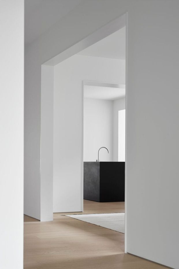 A minimalist white interior viewed through a sequence of open doorways, with pale timber flooring, crisp white walls and ceilings, and a distant black kitchen island with a curved tap. The composition emphasizes clean architectural openings, layered sightlines, and a restrained monochromatic palette.