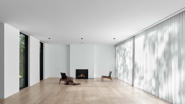 A spacious minimalist living room with pale timber flooring, white walls, a linear recessed fireplace, narrow floor-to-ceiling glazing on the left, and a full-height wall of sheer curtains on the right. Two dark lounge chairs and a low rug provide sparse furnishings, while dappled daylight creates a calm, airy atmosphere.