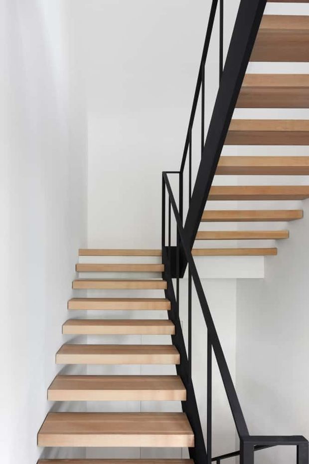 A minimalist contemporary stairway with floating timber treads and a black metal balustrade. The stairs turn at a landing within a bright white interior, creating a strong geometric composition with clean lines and open risers.