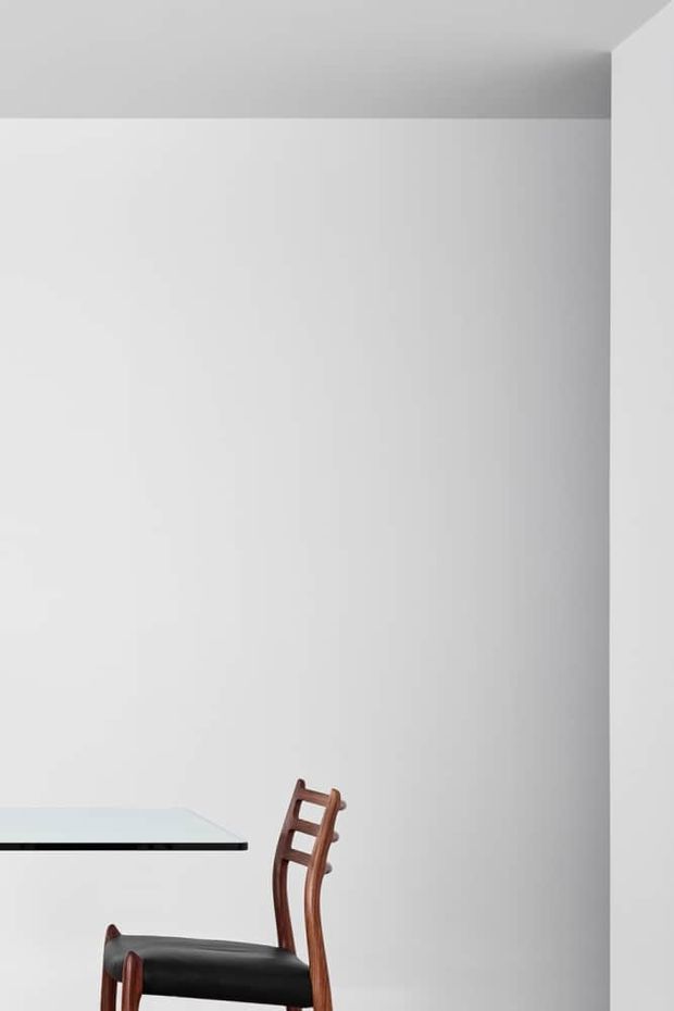 Minimalist interior corner with a largely blank white wall and ceiling, a partial glass-topped table with a dark edge, and part of a dark wooden chair with a black seat. The image is sparsely furnished and brightly lit, with clean architectural lines and neutral tones.