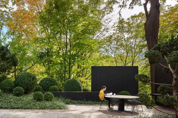 A landscaped outdoor courtyard or terrace set within mature woodland, featuring clipped rounded shrubs, groundcover planting, large established trees, and a minimalist black privacy or feature wall. A small outdoor dining setting with a round table and chairs sits on a paved stone terrace, creating a refined garden entertaining area.