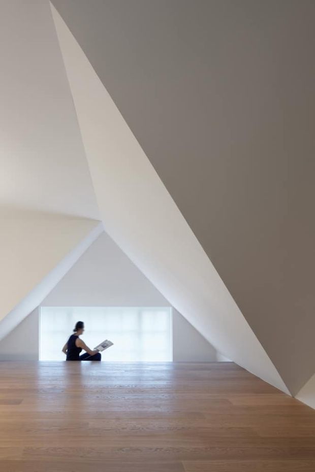 Minimalist interior with a dramatic angular, vaulted ceiling and sculptural white plaster forms. A broad light timber floor leads toward a low, softly diffused window, with a seated person reading near the opening. The space is sparse, airy, and strongly defined by geometric architecture and natural light.