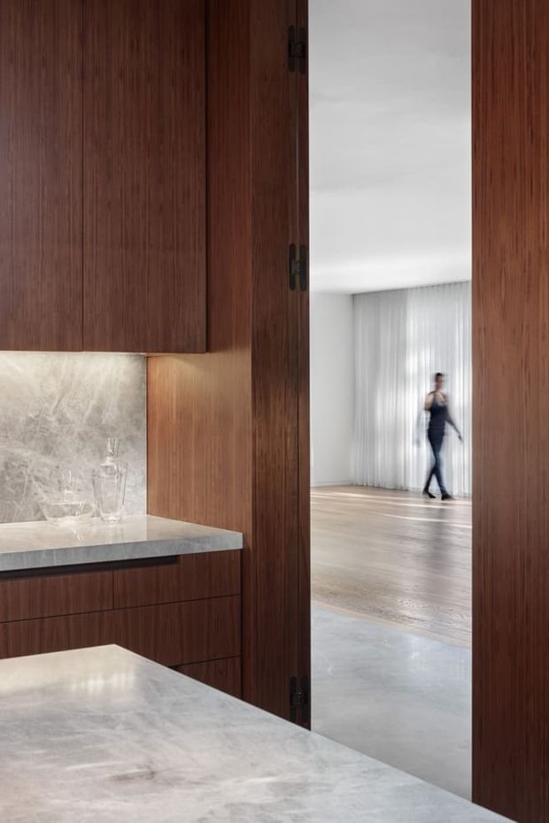 A refined contemporary kitchen or kitchenette viewed through an open, dark timber-framed doorway. The foreground features rich walnut-toned vertical-grain cabinetry, a pale veined stone benchtop and matching stone splashback, with integrated under-cabinet lighting. Beyond the opening is a bright, sparsely furnished living area with full-height sheer curtains and timber flooring; a blurred person is walking in the background.