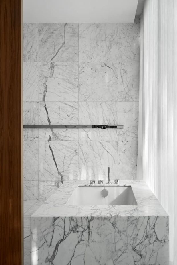 A refined bathroom vanity with a rectangular integrated basin set into a thick white marble countertop and waterfall-front vanity. Matching veined marble tiles cover the wall, with a slim dark horizontal shelf, minimalist chrome tapware, a warm timber door or panel at left, and sheer white curtains at right. The composition is bright, monochromatic, and luxurious.