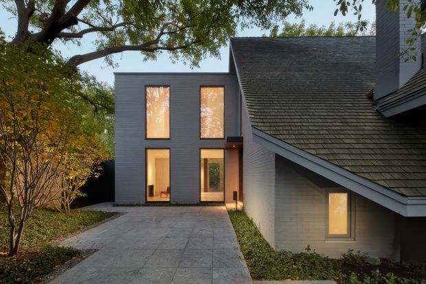 Exterior architectural view of a contemporary multi-volume home at dusk, showing a dark grey horizontally clad two-storey extension with tall illuminated windows beside an older pitched-roof volume. A concrete-paved driveway or walkway leads toward the entrance, with mature trees and landscaped planting surrounding the building.