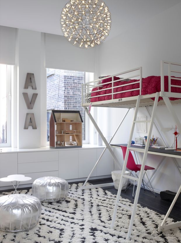 Bright contemporary kids' room with a raised loft bed, integrated study desk, vivid pink bedding and chair, white built-in cabinetry, large windows, and a sculptural spherical pendant light. A black-and-white patterned rug and metallic silver ottomans add graphic and playful accents, while a miniature dollhouse sits on the window ledge.