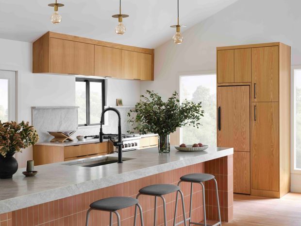 Bright contemporary kitchen with a large island featuring a pale stone benchtop, terracotta vertical tile cladding, and three bar stools. Warm timber cabinetry includes overhead cupboards and a tall integrated pantry unit. Black-framed windows and a dark gooseneck tap contrast with the light walls, while pendant lights and greenery add warmth.