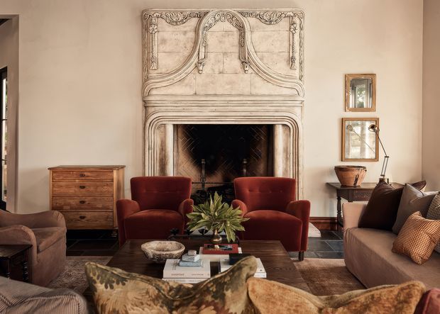 Elegant traditional living room centred on an ornate carved stone fireplace with a dark firebox. The room features deep red velvet armchairs, a large neutral sofa, patterned upholstered seating, a dark timber coffee table, antique-style timber chest, framed mirrors, and warm beige walls with dark timber trim.