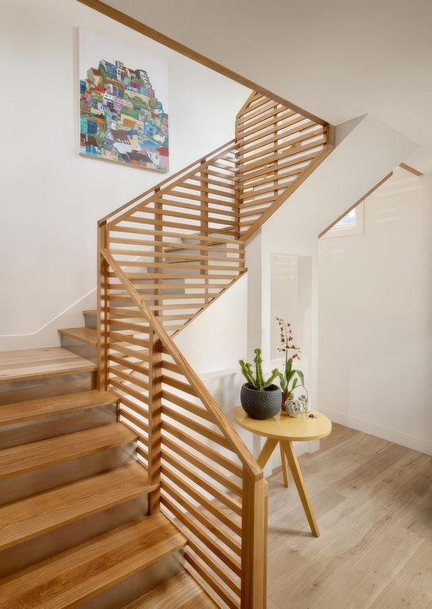 Bright interior view of a timber staircase and landing. The stair has warm wood treads, a slim timber handrail, and an open horizontal-slat balustrade enclosing the stair void. White walls and ceiling, pale timber flooring, a small round timber side table with potted plants, and colourful wall artwork create a clean, warm contemporary setting.