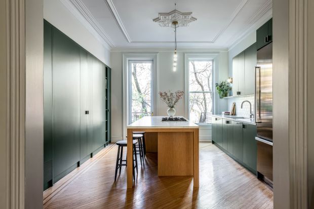 A refined galley-style kitchen viewed from an adjoining room, featuring dark green full-height cabinetry on both sides, a central timber island with seating, white benchtops, stainless-steel appliances, and a warm herringbone timber floor. Tall sash-style windows, ornate ceiling mouldings, and a decorative ceiling medallion bring traditional architectural character to the contemporary fit-out.