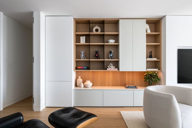 A bright contemporary living room featuring a full-width built-in entertainment and display wall. The joinery combines warm timber open shelving, white cabinetry, vertical timber-look ribbed paneling, and concealed television storage. Light timber flooring, a neutral upholstered armchair, black leather lounge furniture, and restrained decorative objects create a refined, modern interior.