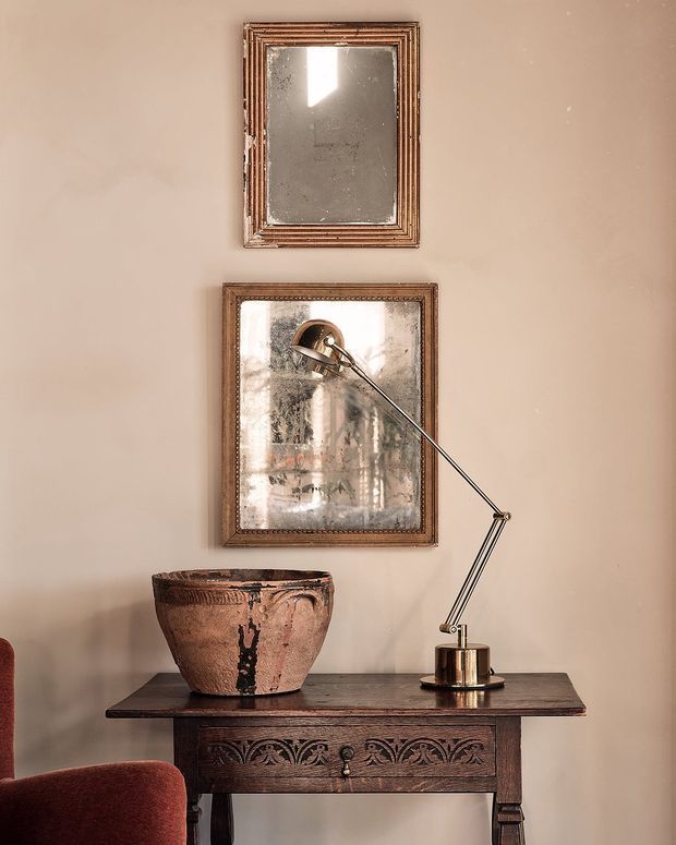 A styled interior vignette featuring an antique dark timber console table with carved drawer detailing, a large weathered terracotta vessel, and a slender articulated metallic desk lamp. Two aged framed mirrors hang above on a warm taupe plaster wall, with part of a rust-coloured upholstered chair visible at the edge. The composition has an eclectic, vintage and refined character with warm, muted tones.