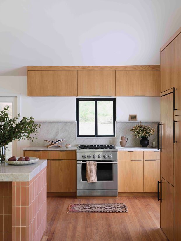Bright, compact kitchen with warm timber cabinetry spanning the rear and right walls, a stainless-steel freestanding range, pale stone benchtops, and a grey stone backsplash. A black-framed window sits above the cooking area, while a peninsula with a pink terracotta-tile face occupies the foreground. Timber flooring, natural decor, and a patterned runner create a warm, refined contemporary look.