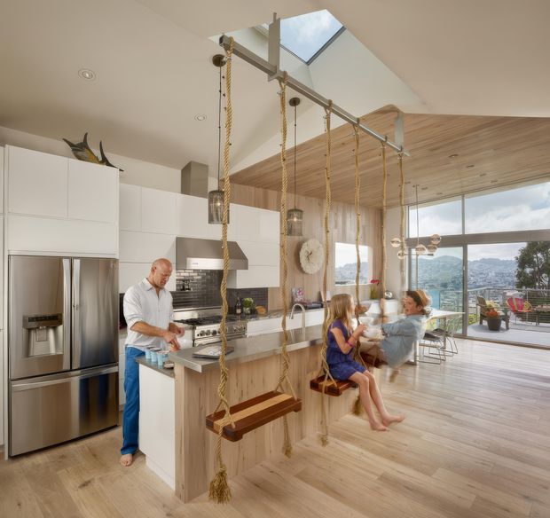 Bright open-plan kitchen and living area with white flat-panel cabinetry, stainless-steel appliances, a timber island and timber-lined ceiling. A dramatic skylight and floor-to-ceiling glazing bring in abundant daylight and reveal a city-and-water view. Rope-hung timber swings form a playful feature beside the island, while a dining and lounge area extends toward the glazed terrace.