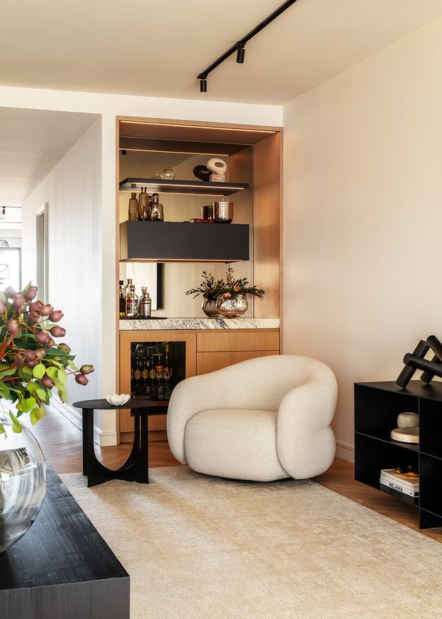 Warm, refined living room with a sculptural cream lounge chair, black side table, black low cabinetry, and a built-in timber bar or display joinery unit with integrated shelving, lighting, bottles, and decorative objects. A large neutral rug, dark timber coffee table edge, track lighting, and a leafy floral arrangement complete the contemporary interior.