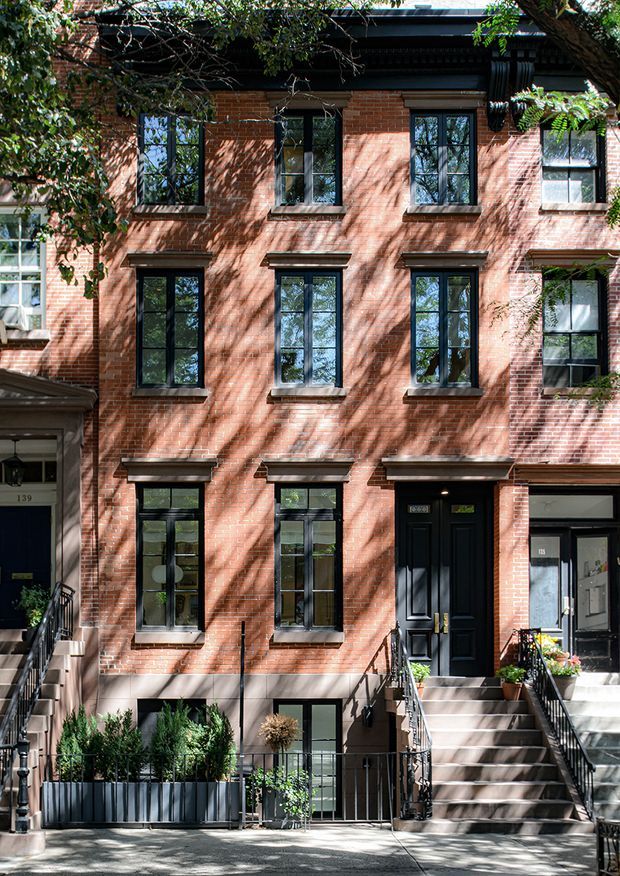 Street-facing facade of a narrow multi-storey brick townhouse or terrace building. The elevation features red brick masonry, regularly spaced tall black-framed windows with projecting stone or concrete sills and lintels, a dark cornice and roof edge, and recessed dark-painted entry doors accessed by raised staircases with black metal railings. Planters line the sidewalk, while mature tree branches cast dappled shadows across the facade.