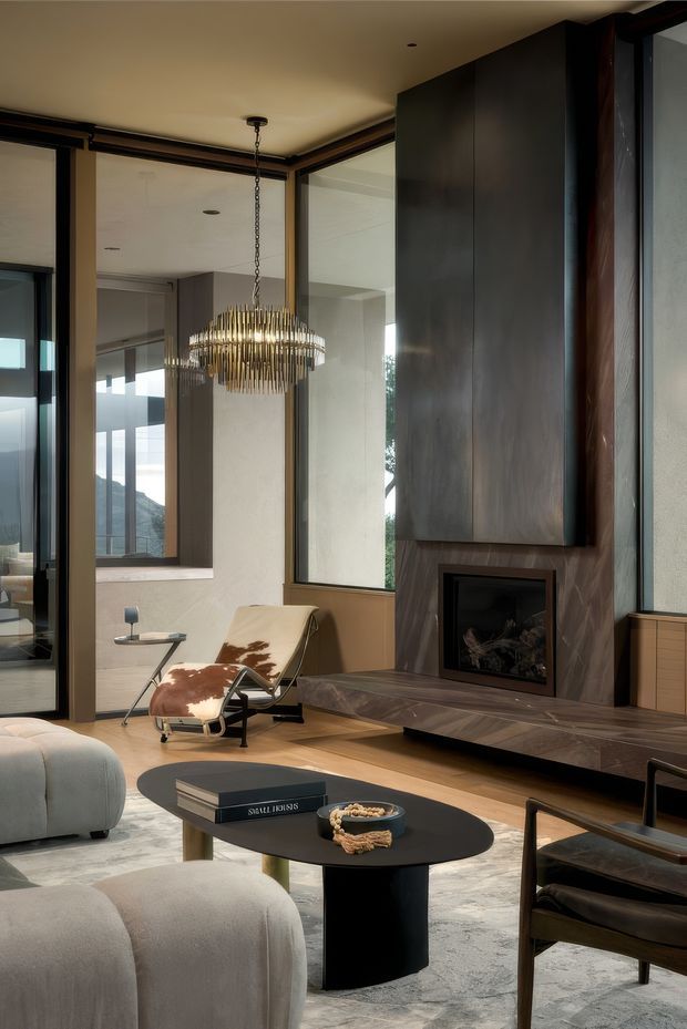A sophisticated contemporary living room with floor-to-ceiling glazing, a dramatic dark stone or metal-clad fireplace wall, and a suspended sculptural chandelier. The room includes a low black oval coffee table, curved upholstered seating, a leather-and-metal lounge chair, and a neutral textured rug. Warm timber flooring and expansive glazing create a refined connection to the outdoors, with a mountain landscape visible beyond.