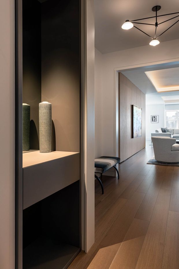 A refined interior view looking from a hallway or entry area into an open living room. The foreground features a tall, dark built-in display niche with integrated shelving and two textured decorative vessels. Warm timber flooring extends through the space, while the living room beyond has neutral upholstered seating, a timber-toned feature wall, artwork, and a contemporary black geometric pendant light. Recessed cove lighting and layered ceiling details add a polished, softly illuminated atmosphere.