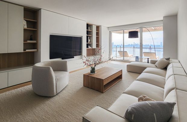 A bright contemporary living room with expansive waterfront-facing glazing. The room features a large cream sectional sofa, rounded upholstered lounge chair, textured neutral rug, timber coffee table, and a wall of integrated cabinetry surrounding a television. Vertical timber slat detailing and open shelving add warmth, while a covered terrace and distant city skyline are visible beyond the glass doors.