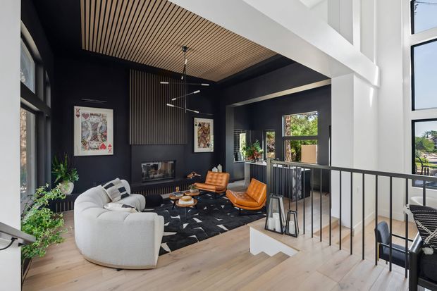 A contemporary double-height living room viewed from an upper-level landing, featuring a curved white sofa, orange leather lounge chairs, a black geometric rug, dark charcoal walls, a linear fireplace, vertical timber slat detailing, and a warm timber slatted ceiling. Black metal balustrading overlooks the space, with large windows providing abundant natural light.