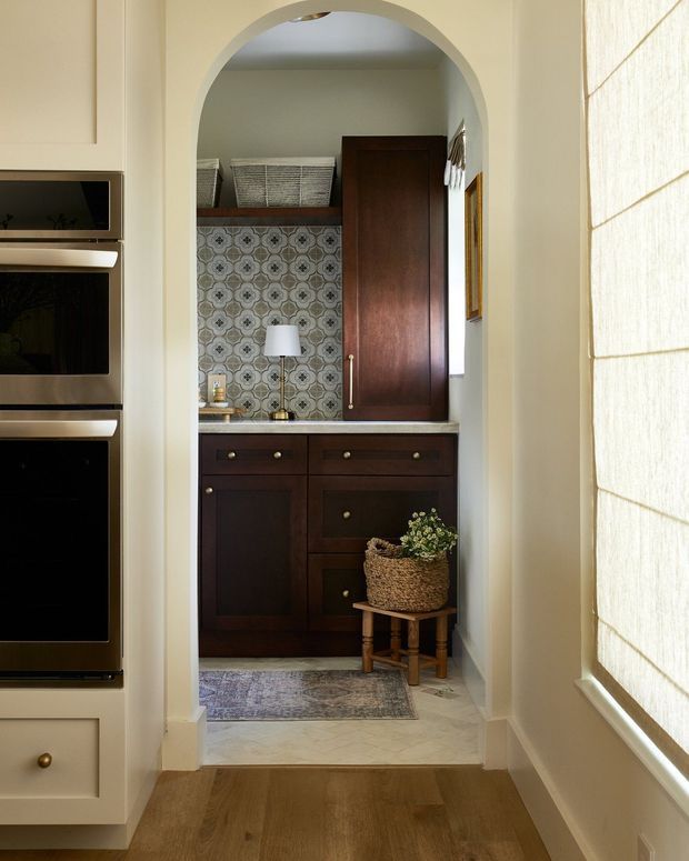 A narrow, elegantly detailed built-in dressing or storage area viewed through a rounded archway. The space features dark wood cabinetry with brass knobs, a patterned tile backsplash, pale marble-look flooring, a woven basket planter, and a small lamp. Cream walls, integrated appliances or cabinetry at left, and a softly filtered window blind at right create a warm, traditional interior with strong contrast between light finishes and dark joinery.