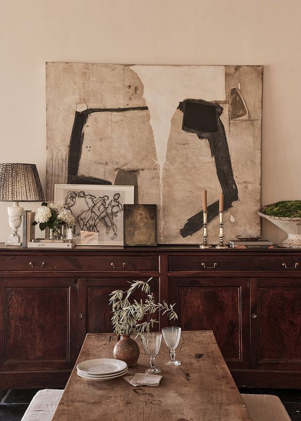 A richly styled traditional dining area featuring a dark stained wood sideboard with panelled cabinetry, a rustic timber dining table, vintage-inspired tableware, floral arrangements, candlesticks, and layered artwork against a warm beige wall. The composition has an elegant, collected, moody character with strong antique and artisanal details.