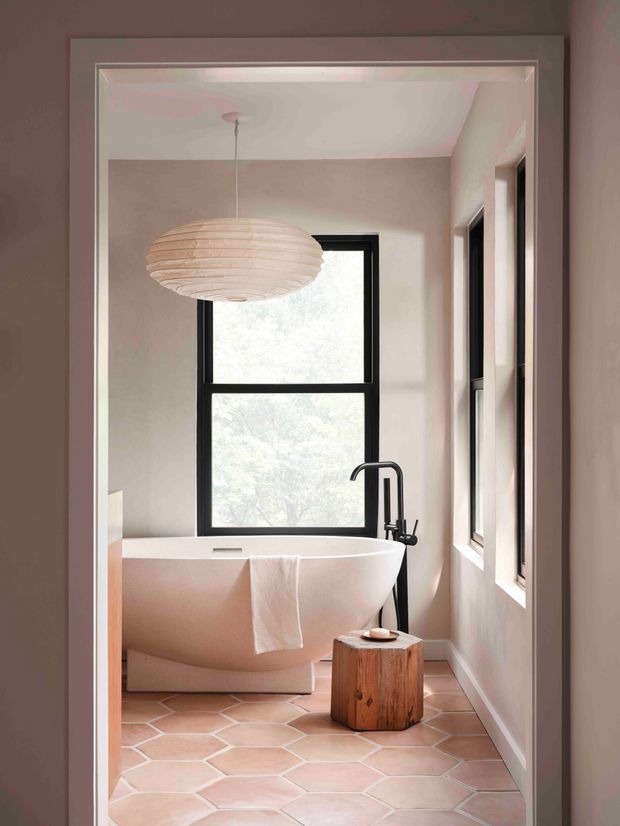 A softly styled bathroom viewed through a doorway, featuring a freestanding white bathtub beneath a large black-framed window, a dark floor-mounted bath tap, pale pink plaster walls, terracotta-toned hexagonal floor tiles, and a sculptural wood stool. A large ribbed pendant light hangs above the bath, creating a warm, serene atmosphere.