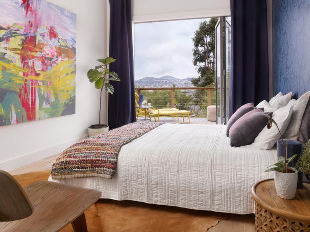 A stylish bedroom with a large white quilted bed, layered pillows, a colourful woven throw, dark navy curtains, and a blue patterned feature wall. An abstract artwork and indoor plant add colour, while full-height glazing opens to a balcony with outdoor seating and views toward distant hills. Warm timber flooring and bedside furniture create a comfortable, refined atmosphere.