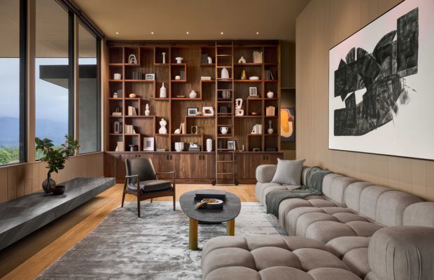 A refined contemporary living room with floor-to-ceiling corner glazing, a built-in dark timber shelving and display wall, a long sculptural modular sofa, lounge chair, round coffee table, and large abstract artwork. Warm timber wall panelling and flooring contrast with soft grey upholstery and a textured rug, while expansive windows provide a distant landscape view.