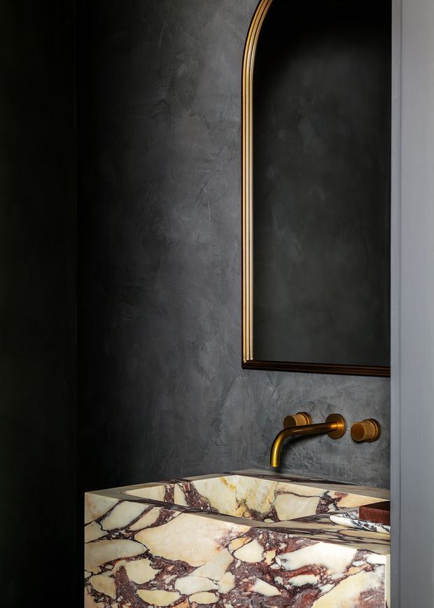 A compact, dark-toned bathroom vignette featuring a sculptural marble basin and integrated backsplash in heavily veined cream, burgundy, and brown stone. The walls have a textured charcoal plaster finish, complemented by a wall-mounted brushed brass tap and matching controls. A tall rounded-arch mirror with a dark brass frame rises above the vanity, creating a refined, dramatic and luxurious atmosphere.