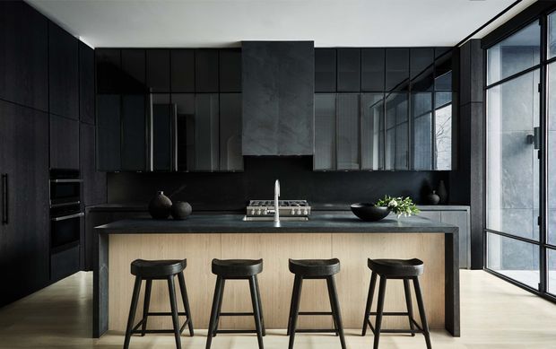 A sophisticated contemporary kitchen with dark floor-to-ceiling cabinetry, a dark stone-look splashback and benchtop, and a large light timber island with four black bar stools. Full-height black-framed glazing along the right side brings in natural light, while integrated appliances and a broad reflective splashback reinforce the sleek, minimal design.