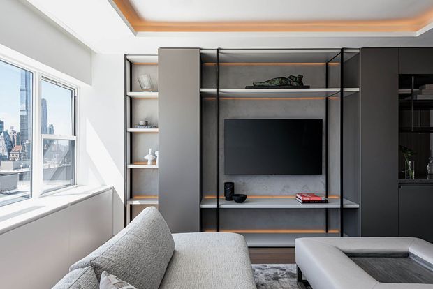 Bright contemporary living room with a large sectional sofa facing a built-in black and charcoal media wall. The wall features open shelving, closed cabinetry, a wall-mounted television, subtle warm wood-toned shelf and ceiling accents, and decorative objects. Large corner windows provide expansive city views and strong natural light.
