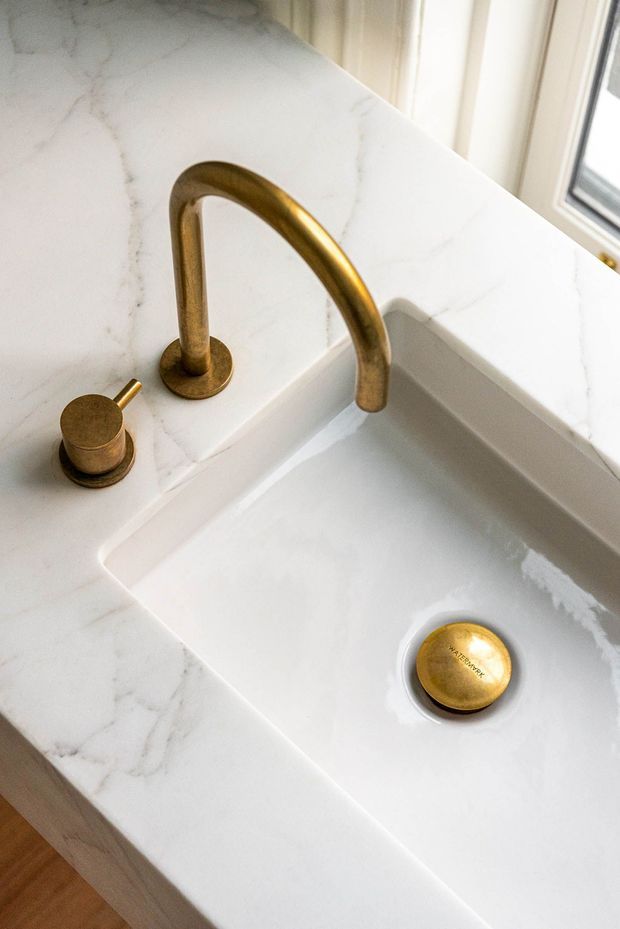 Close-up view of a bathroom vanity or washbasin with an integrated white basin, white veined stone countertop and matching raised backsplash. The basin has a curved brushed brass/gold faucet, separate brass control, and brass drain cover. A window is visible at the upper right, with light timber cabinetry or flooring below the counter.