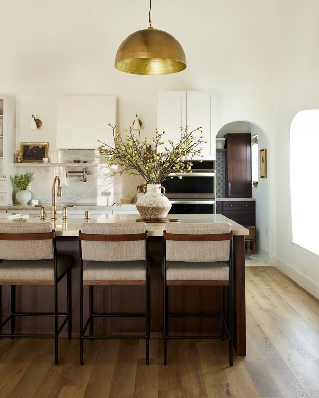 A bright, refined kitchen centered on a large island with a pale stone benchtop, dark timber cabinetry, and three upholstered bar stools. White paneled cabinetry, integrated appliances, a marble-look splashback, brass tapware, and a large aged-brass pendant define the space. Light timber flooring and an arched doorway add warmth and architectural character.