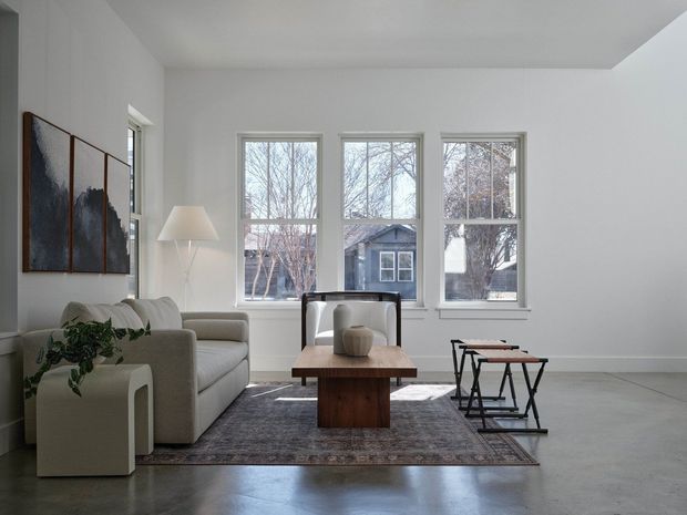 Bright, minimalist living room with high ceilings, large multi-pane windows, a pale upholstered sofa, sculptural armchair, low timber coffee table, floor lamp, abstract wall art, and nesting side tables. The room has a polished grey concrete floor, white walls, and a muted grey-brown area rug, with a leafy plant adding a small natural accent. Residential buildings and trees are visible outside the windows.