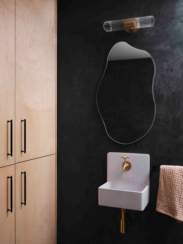 A compact, dramatically styled bathroom vanity area with a black textured wall, an organically shaped frameless mirror, a sculptural white wall-mounted basin, and a brass tap and exposed plumbing. Pale timber cabinetry with slim black handles occupies the left side, while a textured beige towel hangs to the right. A linear ribbed wall light with brass detailing sits above the mirror.