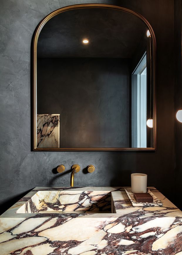 A dark, sophisticated bathroom vanity featuring a large arched mirror with a thin metallic frame, charcoal textured wall finish, warm wall and ceiling lighting, and a sculptural brass wall-mounted tap. The vanity, integrated basin, and surrounding benchtop are clad in richly veined cream and burgundy natural stone. A narrow window is visible to the right, while a cup and small accessories sit on the countertop.