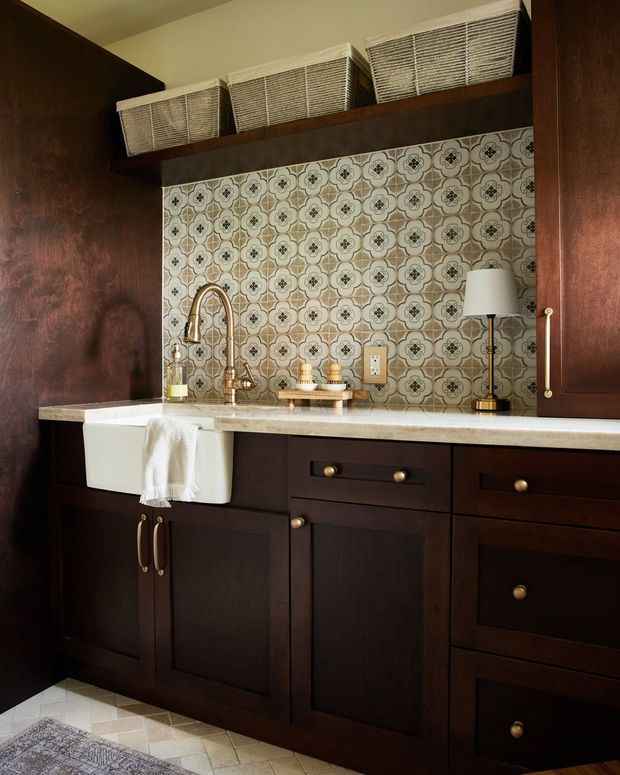 A compact, richly detailed kitchen or butler's pantry with dark stained wood cabinetry, a white farmhouse sink, brass fixtures and hardware, a pale stone benchtop, and an ornate patterned tile splashback. Woven baskets sit on an open shelf above the splashback, with a small table lamp adding warmth.