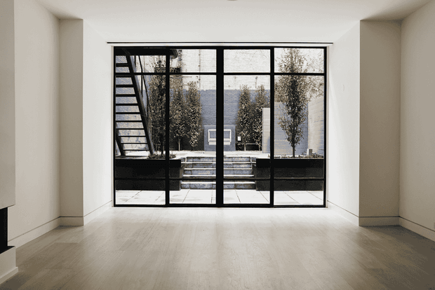 Minimal, light-filled interior room with pale timber flooring and smooth off-white walls, focused on a large black-framed glazed opening. Through the glazing is a stepped courtyard with planters and trees, while a slim black staircase is visible at the left. The space is largely unfurnished and has a calm, contemporary architectural character.